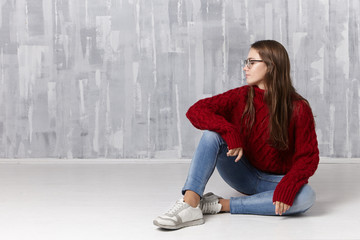 Beauty, style, fashion, youth, people and lifestyle concept. Studio shot of nice charming teenage girl with long loose hair sitting on floor, wearing glasses, turtleneck sweater, jeans and sneakers