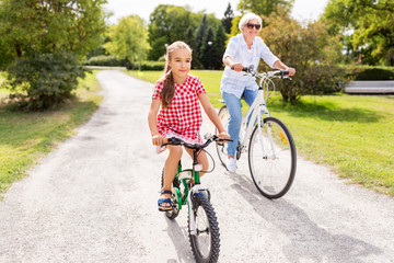 family, leisure and people concept - happy grandmother and granddaughter riding bicycles at summer...