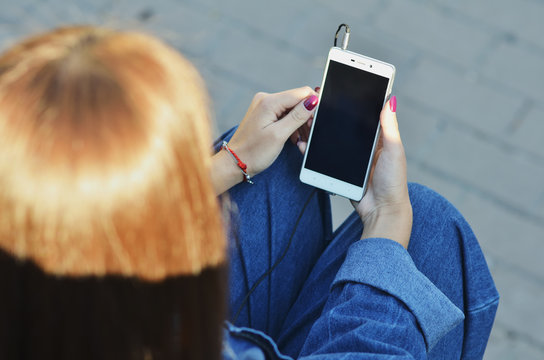 Smartphone close-up in the hands of a girl dressed in denim clothing that listens to music in headphones