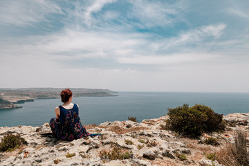 Fototapeta na wymiar Young redhead woman with a topknot sitting on the edge of a cliff looking far away the sea