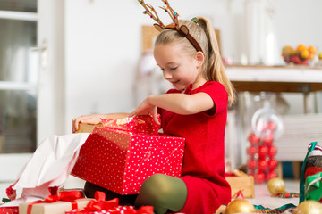 Cute super excited young girl opening large red christmas present while sitting on living room...