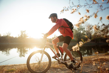 Determined young bearded man in long sleeve jersey riding a mountain bike by the lake or river. Sunset over water in background. Side view