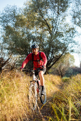 Smiling young bearded man in red long sleeve cycling jersey riding mountain bike along a path through tall grass. Low angle front view.