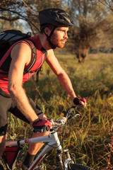 Determined young bearded man in red sleeveless cycling jersey riding mountain bike along a path through forest. Standing on the pedals and sprinting
