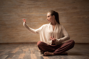 Calm young woman putting her hand sideward and looking into the distance while sitting in the lotus...