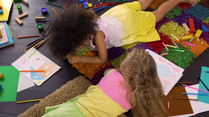 Adorable curly multiracial friends lying on the floor and gossiping at art club