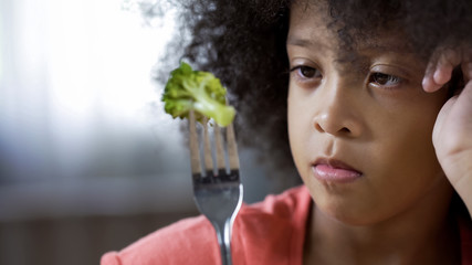 Adorable Afro-American girl sadly looking at fork with broccoli piece, eco food