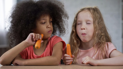 Cute little girl looking at friend eating carrots with appetite, healthy food