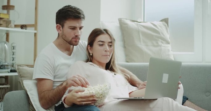Weekend time for a young couple spending a good time together in living room sitting on the sofa eating popcorn and watching movie from notebook.
