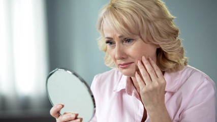 Sad woman looking at face reflection and crying, unhappy with wrinkled skin