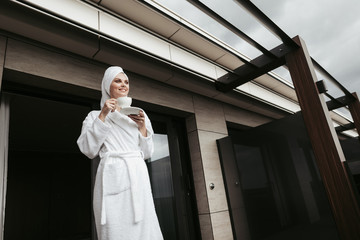 Concept of relaxation and harmony after spa. Full length low angle portrait of young beautiful lady in bathrobe drinking tea with pleasuring smile on terrace. Copy space on right