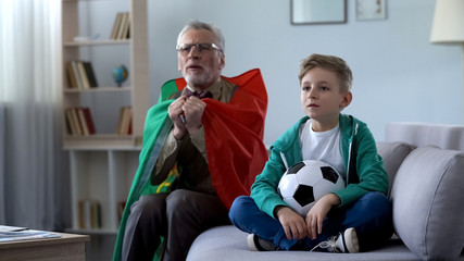 Grandpa wrapped in Portuguese flag watching soccer with boy, worrying about game