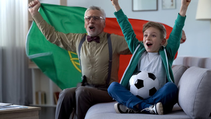 Grandpa and grandson watching football, waving Portuguese flag, happy for win