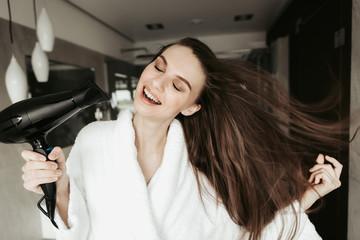 Fototapeta na wymiar Concept of haircare and enjoyment after shower. Close up portrait of cheerful beautiful woman closing eyes while drying hair with blowdryer at home bathroom interior