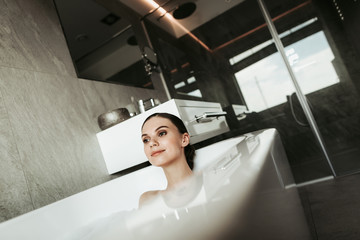 Concept of water enjoyment and relaxation at home. Close up portrait of beautiful brunette woman with charming smile lying in relaxing home bath with foam
