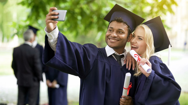 Young people in graduation outfit taking selfie with mobile phone, looking at it