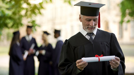 Senior man in academic regalia holding diploma, education at any age, new degree