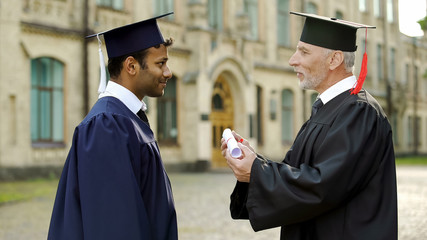 Qualified professor giving diploma to mixed-race male student congratulating him - obrazy, fototapety, plakaty
