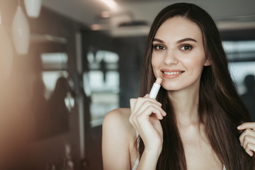 Portrait of happy lady with attractive smile rouging lips while locating in room. Beauty concept