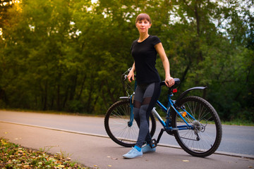 Obraz premium Young beautiful athletic girl in sportswear sitting on a blue bike frame. Trees on the background.
