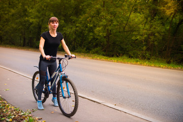 Fototapeta premium Young beautiful athletic girl in sportswear sitting on a blue bike on the sidewalk. Trees on the background.