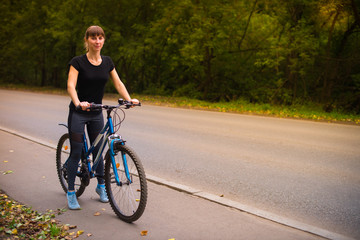 Young beautiful athletic girl in sportswear sitting on a blue bike on the sidewalk. Trees on the background.