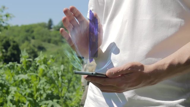 Man shows concept hologram Adapt on his phone. Person in white t-shirt with future technology holographic screen and green nature background