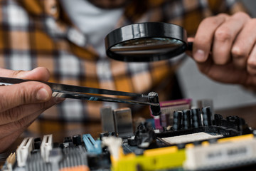 cropped shot of computer engineer with tweezers and magnifying glass repairing motherboard