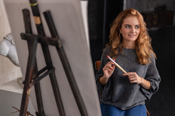 Woman painter holding a brush and palette with oil paints in her hand, standing in front of an empty canvas, drawing.