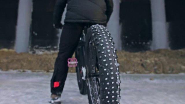 Professional extreme sportsman biker riding fat bike in outdoors. Cyclist ride in winter on snow ice under bridge. Man do trick on mountain bicycle with big tire in helmet glasses. Slow motion 180fps.