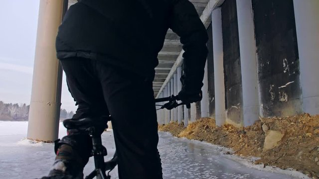 Professional extreme sportsman biker riding fat bike in outdoors. Cyclist ride in winter on snow ice under bridge. Man do trick on mountain bicycle with big tire in helmet glasses. Slow motion 180fps.
