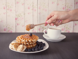 Woman pouring honey on delicious waffles with bananas and blueberries