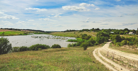 Sunny summer landscape with ground country road,river and clouds in blue sky.