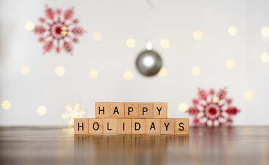 2018 Seasonal Festive Photo Banner Idea of Happy Holidays Spelled Out of Stacked Wood Block Letters with Colorful Red Snowflake Ornaments and Bright Yellow Twinkle Lights in Blurred White Background 