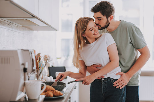 Young bearded man gently kissing his charming girlfriend while she stirring something in pan