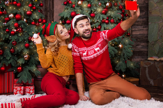 Happy family couple in love with santa and elf hats warm sweaters doing selfie with smartphone for greeting parents with Merry Christmas and Happy 2019 New Year at home with decorated christmas tree