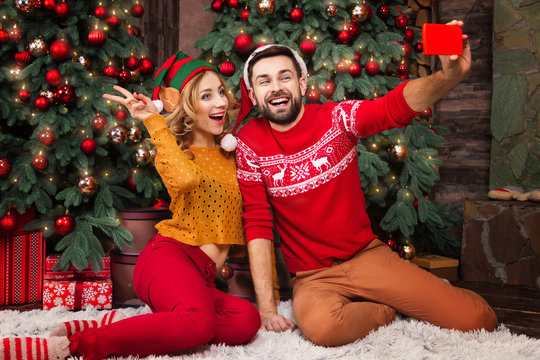 Happy family couple in love with santa and elf hats warm sweaters doing selfie with smartphone for greeting parents with Merry Christmas and Happy 2019 New Year at home with decorated christmas tree