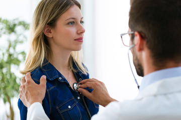 Handsome young male doctor checking beautiful young woman patient heartbeat using stethoscope in...