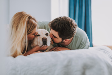 Pleasant young couple resting in bed while kissing their dog