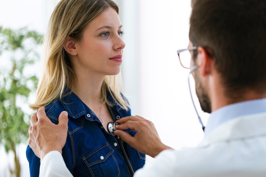 Handsome young male doctor checking beautiful young woman patient heartbeat using stethoscope in medical office.