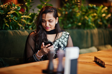 Pretty indian girl in black saree dress posed at restaurant with mobile phone at hand.