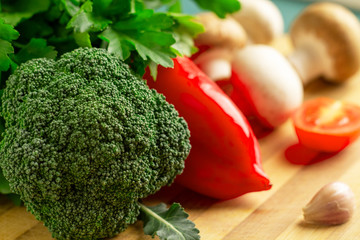Still life of broccoli, pepper, tomato, champignons, royal champignons, garlic, parsley lies on a wooden kitchen board. Focus on broccoli inflorescence.