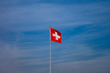 Swiss flag waving on the blue sky on a white mast and a sunny day with clouds in Switzerland.