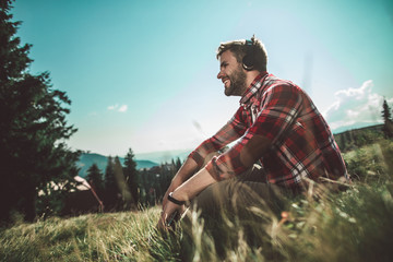 Relaxing in travel with good sounds. Side on low angle portrait of bearded cheerful male sitting on green mountain hill and listening music by mobile phone