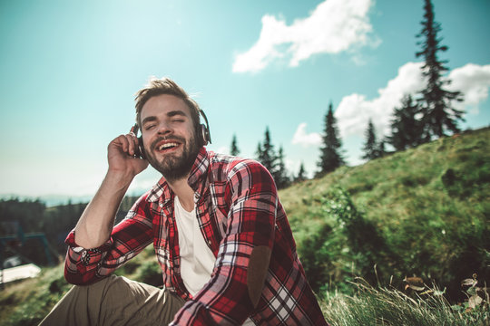 Relaxing and enjoying good sounds on nature. Waist up portrait of bearded cheerful male sitting on green mountain hill and listening music. Copy space on right
