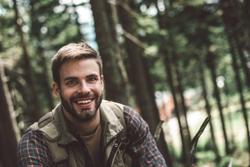Naklejka na ściany i meble Concept of interesting journey and adventure. Close up portrait of smiling young male among green trees cheerfully looking straight on camera. Copy space on right