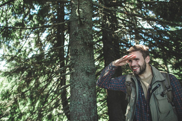 Concept of interesting journey and adventure. Waist up portrait of smiling young male putting his hand on forehead and trying to see something among green trees