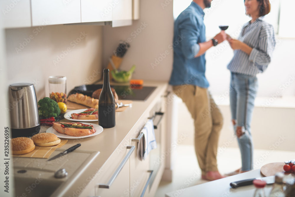 Wall mural Stove, bottle of wine, beef steaks and burger buns in cozy kitchen. Bearded man and his girlfriend enjoying drinks