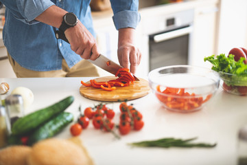 Cooking dinner at home. Close up of male hands slicing red bell pepper with knife on cutting board