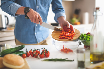 Cooking dinner. Cropped portrait of gentleman in denim shirt holding knife and cutting board with bell pepper while standing near kitchen table
